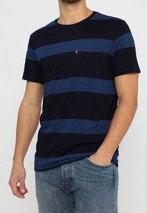 Mężczyzna w granatowo-ciemnoniebieskiej koszulce w poziome paski z krótkim rękawem i kieszonką na piersi, w połączeniu z jasnoniebieskimi dżinsami.