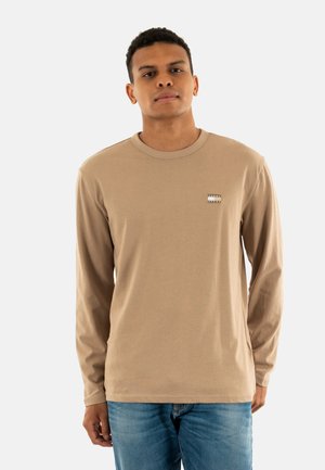 Camicia a maniche lunghe beige in cotone con colletto tondo e una piccola patch con logo sul lato sinistro del petto, abbinata a jeans in denim blu.