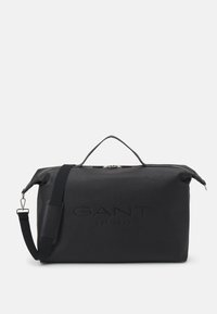 Musta duffel-laukku, joka on valmistettu teksturoidusta synteettisestä materiaalista, varustettu ylhäältä avautuvalla vetoketjulla, kahvalla ja säädettävällä hihnalla. Etupuolella painettu brändilogo.