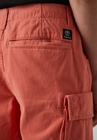 Koralo spalvos krovinių šortai su lygiu paviršiumi, turintys užpakalinį kišenę ir Timberland logotipo etiketę ant juosmens.