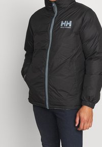 Czarna ocieplana kurtka z wysokim kołnierzem, przednim zamkiem błyskawicznym i elastycznymi mankietami. Na piersi znajduje się jasno niebieskie logo Helly Hansen.