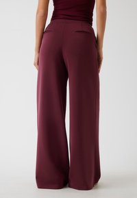 Vrouw in hoge taille, brede burgundy broek staande tegen een effen achtergrond, achteraanzicht.