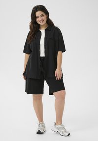 Chemise noire à manches courtes avec deux poches poitrine, associée à un short noir. Des chaussures de sport sont visibles. Design simple et décontracté avec une coupe ample.