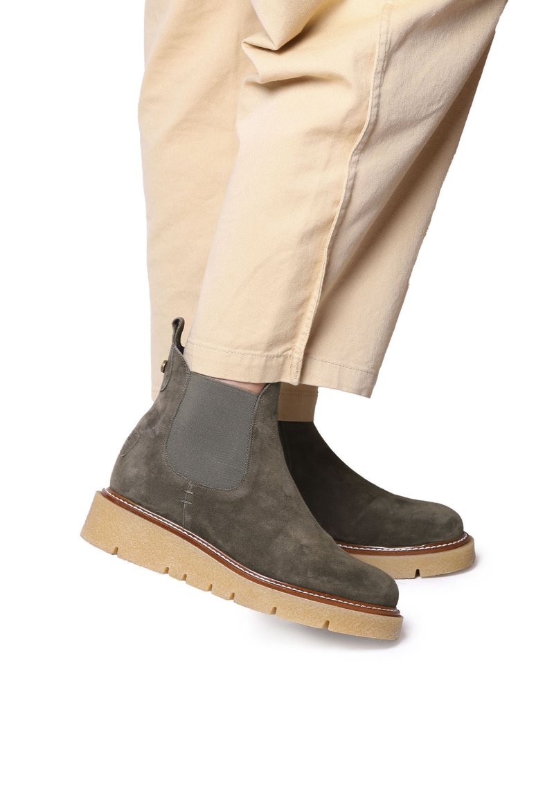 Bottines en daim vert olive avec des panneaux élastiques sur les côtés, semelle en caoutchouc et coutures beige contrastantes. Posées sur un pantalon de couleur claire.