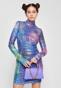 Sukienka z cekinami o wysokim dekolcie; wielokolorowe odcienie niebieskiego, zielonego i różowego; dopasowany fason. Trzyma fioletową torebkę na złotym łańcuszku.
