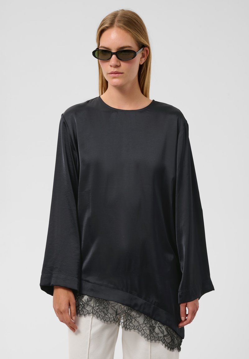 Femme portant un haut en satin noir à manches longues avec un détail asymétrique en dentelle à l'ourlet et un pantalon blanc, portant des lunettes de soleil ovales foncées.