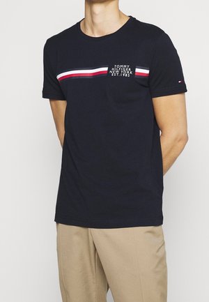 Mężczyzna w granatowej koszulce Tommy Hilfiger z czerwono-biało-granatowymi poziomymi paskami i beżowych spodniach na jednolitym tle.