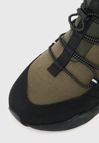 Black and olive green sneaker with textured fabric, black rubber sole, and elastic laces. Features stitched detailing and plastic accents.