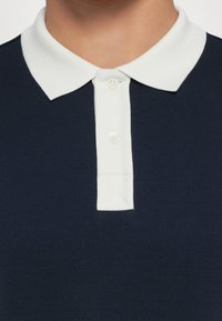 Marineblaues Poloshirt mit einem kontrastierenden weißen Kragen, ausgestattet mit zwei weißen Knöpfen und einem strukturierten Stoff. Zeigt einen dreieckigen Akzent unter dem Kragen.