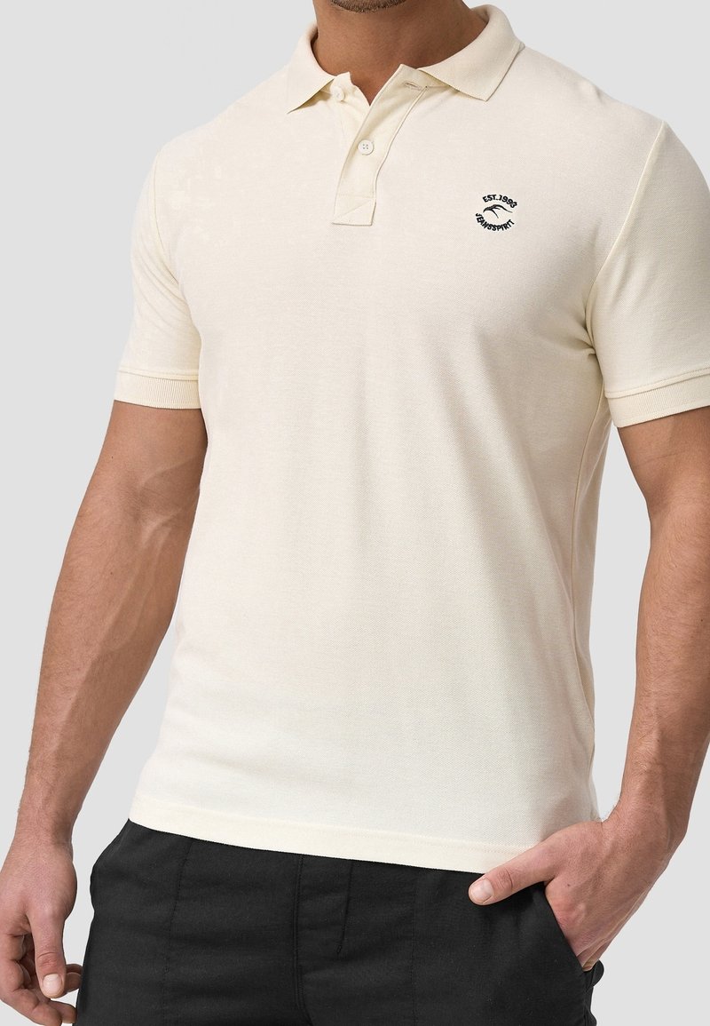 Homme portant un polo crème à manches courtes avec deux boutons et un petit logo noir brodé sur la poitrine gauche.
