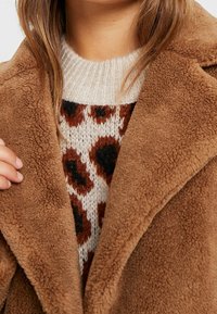 Mujer vestida con un abrigo de teddy marrón sobre un suéter blanco con estampado de leopardo marrón y negro, sujetando la solapa del abrigo con una mano.