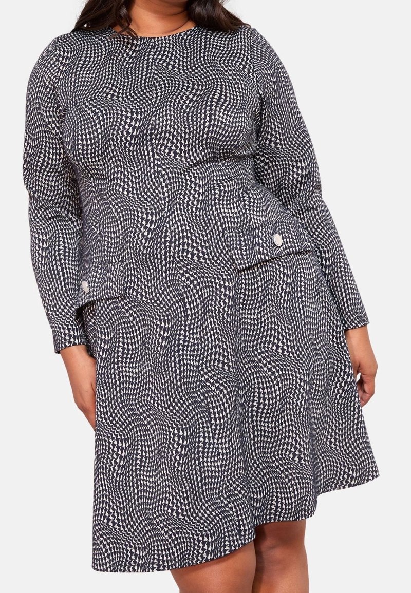 Femme portant un haut à manches longues noir et blanc à motif et une jupe assortie avec des boutons sur les côtés, debout devant un fond uni.