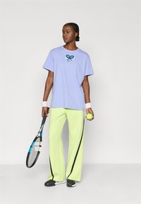 Μωβ ανοιχτό μπλουζάκι με γραφικό σχέδιο ρακέτας τένις, συνδυασμένο με φαρδιά παντελόνια σε φωτεινό lime πράσινο με μαύρες ρίγες. Κρατάει ρακέτα και μπάλα τένις.