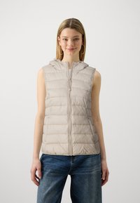 Beige zateplená vesta s předním zipem, bez rukávů a kapucí. Disponuje prošívanou strukturou a přiléhavým pasem, kombinována s modrými džíny.