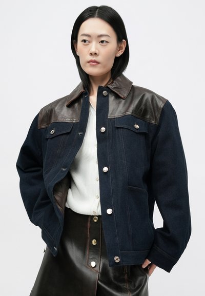 Veste en denim avec des accents en cuir marron aux épaules, couleur bleue, fermetures à boutons et poches latérales. Haut blanc côtelé en dessous.