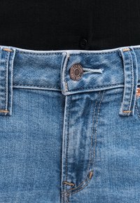 Κοντινό πλάνο της μέσης μπλε τζιν με χάλκινο κουμπί και θηλιές ζώνης πάνω από μαύρο ριμπ ύφασμα.