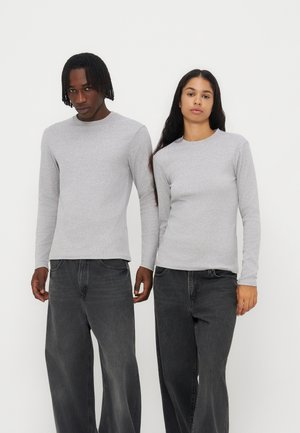 Deux personnes debout côte à côte portant des chemises à manches longues gris clair et des jeans noirs à jambes larges sur un fond blanc uni.