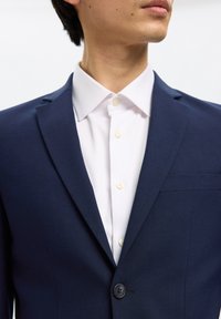 Man die een donkerblauw colbert draagt over een wit overhemd, met de bovenste knoop van het colbert dicht en de kraag open.
