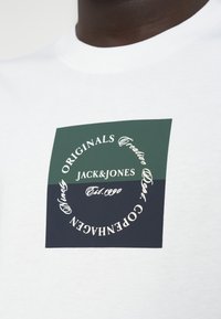 Λευκό βαμβακερό μπλουζάκι με πολύχρωμο ορθογώνιο λογότυπο. Το λογότυπο περιλαμβάνει πράσινα και ναυτικά τμήματα με λευκό κείμενο.