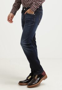 Mörkblå denimjeans med slim fit, som har kontrastsömmar och detaljerade sidofickor, tillsammans med bruna och vinröda läderskor.