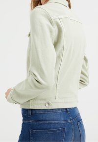 Vihreä denimi takki, jossa on nappikoristeet, leveä kaulus ja istuva siluetti. Näkyvät ompeleet saumoissa ja hihansuiden yksityiskohdissa.