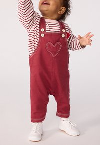 Kleinkind in roten Latzhosen mit gesticktem Herzen, gestreiftem Langarmshirt und weißen Turnschuhen, das nach oben greift.
