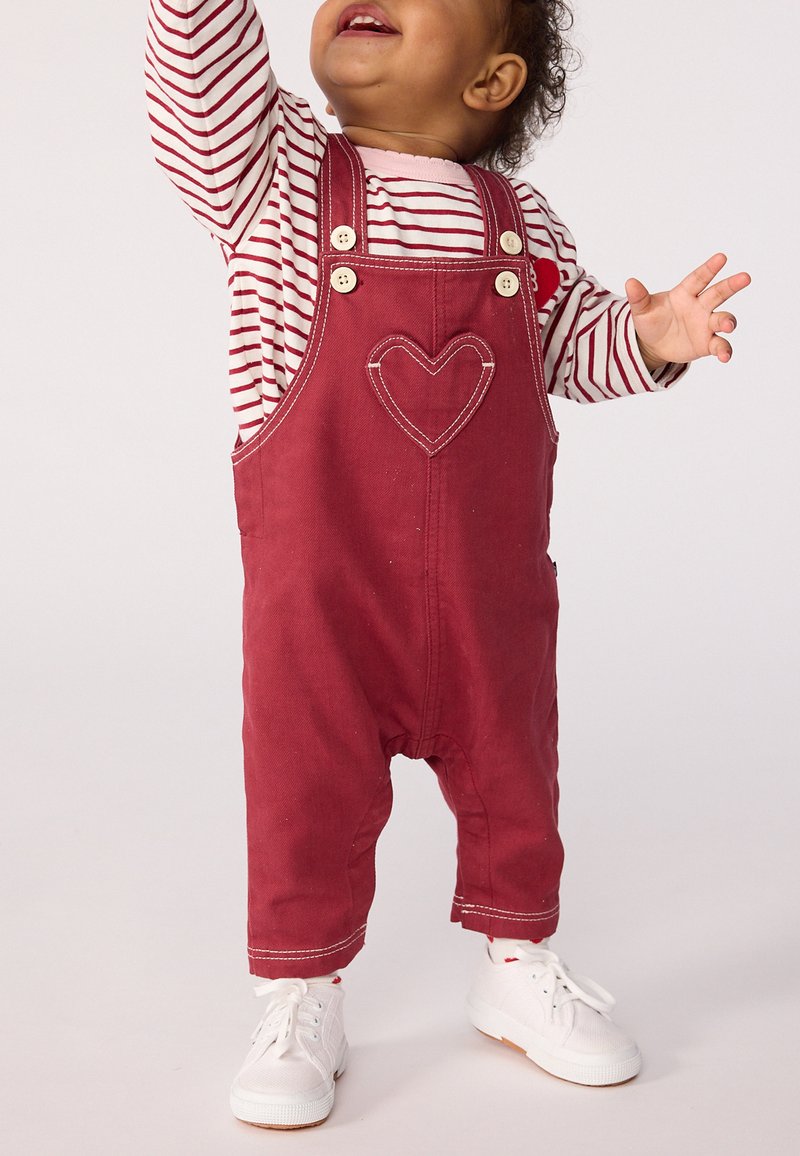 Kleinkind in roten Latzhosen mit gesticktem Herzen, gestreiftem Langarmshirt und weißen Turnschuhen, das nach oben greift.