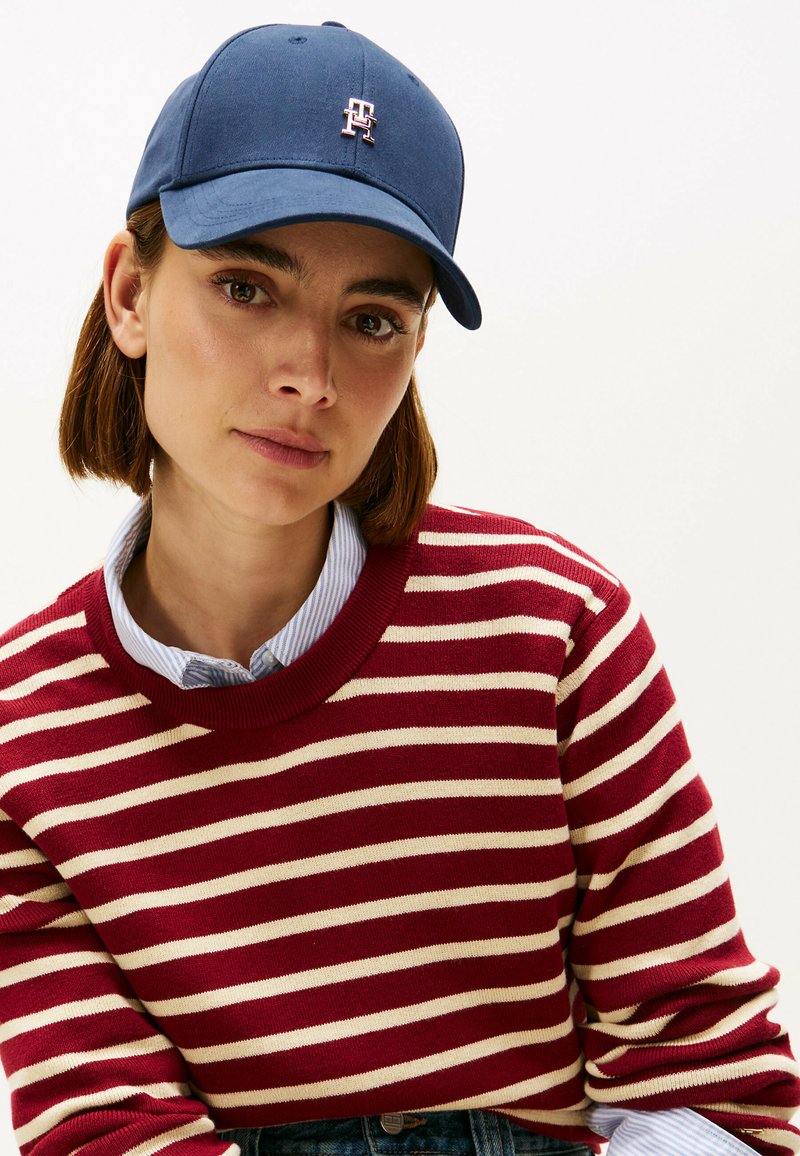 Cappello da baseball navy con logo ricamato, abbinato a un maglione a strisce rosse. Il maglione presenta strisce orizzontali crema e un collo a girocollo.