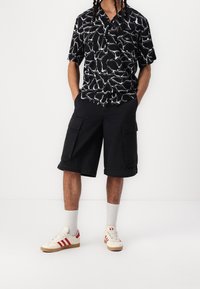 Černé tričko s krátkým rukávem a bílým abstraktním vzorem, kombinované s černými cargo šortkami se bočními kapsami. Bílé a červené tenisky se šedými akcenty.