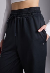 Crne atletske hlače od mekanog materijala, s elastičnim pojasom s vezicom i bočnim džepovima. Diskretan logo na nozi.