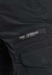 Pantalones cargo negros con una tela texturizada, un bolsillo de parche con costuras decorativas y una etiqueta de marca que dice "PME LEGEND AMERICAN CLASSIC."