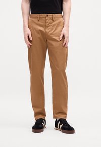 Mand iført slim-fit brune bukser med et sort bælte, sorte og beige sneakers, står med armene afslappet langs siden.
