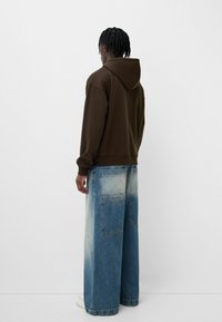 Felpa marrone con una grande tasca posteriore, abbinata a pantaloni in denim blu sbiadito a gamba larga con più tasche e un effetto gradiente.