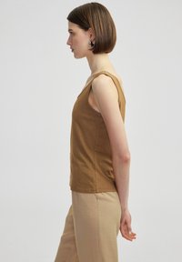 Canottiera marrone senza maniche in un tessuto morbido, caratterizzata da una vestibilità comoda e scollatura rotonda, abbinata a pantaloni beige chiari.