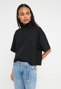 Frau, die ein schwarzes kurzärmliges Rollkragenoberteil und hellblaue, hoch taillierte Jeans mit einem passenden Stoffgürtel trägt, vor einem weißen Hintergrund steht.