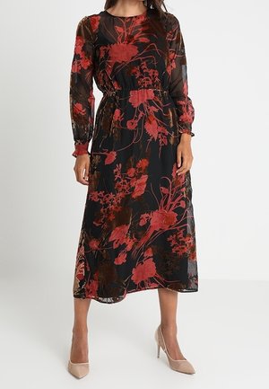 Zwarte bloemenjurk met lange doorzichtige mouwen, een cinched taille en een vloeiende silhouet met rode en bruine bloemenpatronen. Gecombineerd met neutrale hakken.