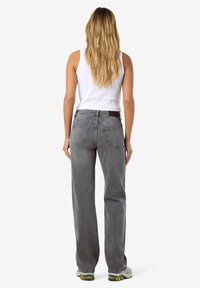 Femeie cu păr lung și blond, purtând un top alb fără mâneci, blugi gri cu talie înaltă și croială dreaptă, și adidași albi, stând cu fața spre departe.