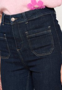 Vaqueros ajustados de mezclilla azul oscuro con cintura alta, cinco bolsillos y detalles de costura visibles. La tela parece elástica y suave.