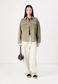 Veste en velours côtelé vert olive avec deux poches poitrine, boutons en argent ; portée sur une chemise noire et associée à un pantalon ample crème et des chaussures noires.