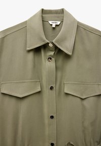 Camicia verde oliva con colletto classico, caratterizzata da bottoni a pressione argentati e due tasche sul petto, realizzata in tessuto liscio con un leggero lucido.