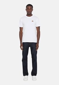 T-shirt bianco a maniche corte con un piccolo logo colorato sul petto, abbinato a jeans blu scuro e sneakers bianche.