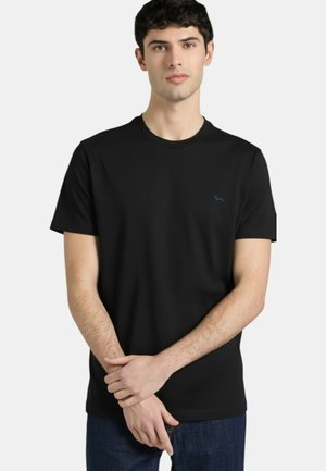 Giovane uomo con capelli corti e scuri che indossa una semplice t-shirt nera a maniche corte e jeans blu scuro, in piedi con le mani giunte.