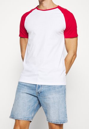 Mężczyzna w białej koszulce z krótkim rękawem i czerwonymi rękawami raglanowymi, w jasnoniebieskich szortach dżinsowych, stoi z rękami za plecami.