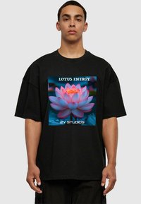Černé oversized tričko s barevným grafikou lotosu, s nápisy "LOTUS ENERGY" a "2Y STUDIOS" v bílém, které jsou výrazně zobrazeny.