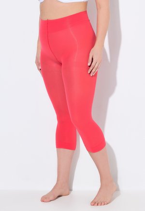 Frau trägt knallpinke, eng anliegende Capri-Leggings, steht barfuß vor einer schlichten weißen Wand, oberhalb der Taille beschnitten.
