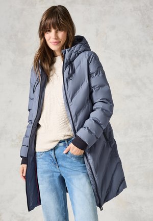 Abrigo gris acolchado con capucha, cremallera frontal y bolsillos laterales. Lleva puesto un suéter cremoso y jeans azules, mostrando un diseño aerodinámico.