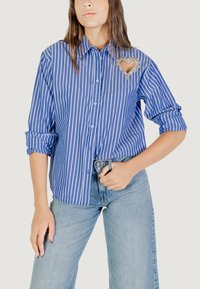 Camicia a righe blu e bianche con decorazione a forma di cuore, maniche arrotolate e vestibilità rilassata. Indossata con jeans azzurri.