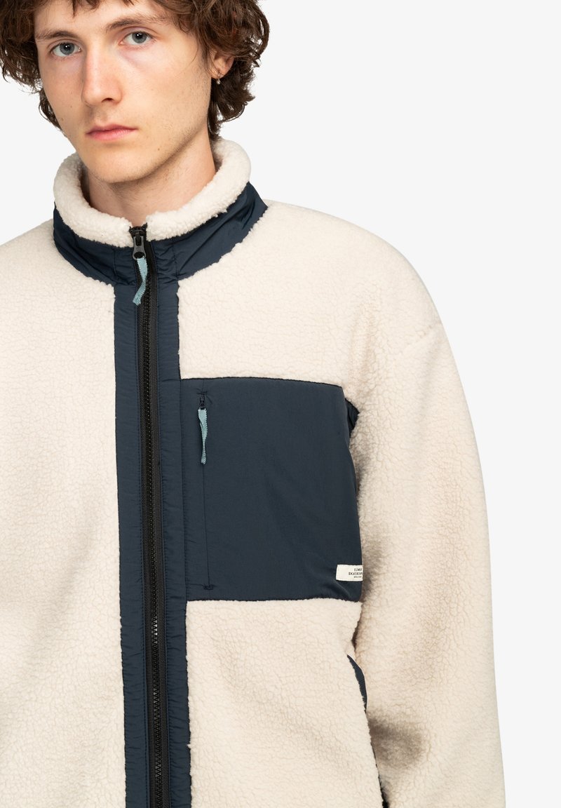 Chaqueta de forro polar con un cuerpo color crema y acentos en azul marino, que presenta un cuello alto, un bolsillo en el pecho con cremallera y una tela texturizada para mayor calidez.