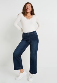 Jeune femme avec de longs cheveux portant un haut blanc à bordure en dentelle, un jean large noir et des baskets blanches, posant sur fond blanc uni.