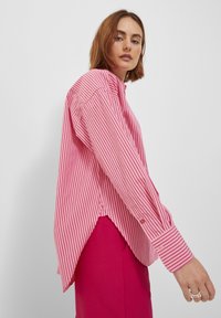 Pink and white striped shirt with long sleeves, oversized fit, and high-low hem. Features button details on sleeves and side slits.
